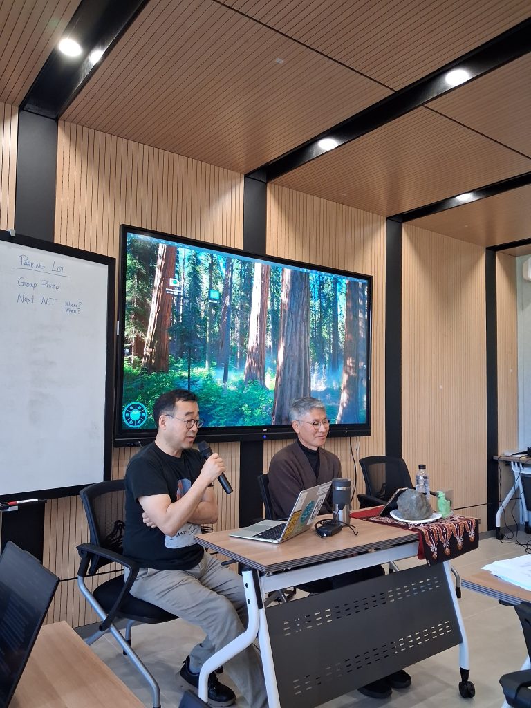 Two people sit at a table with laptops and microphones. A large screen displays a forest scene. A whiteboard with notes is on the left.